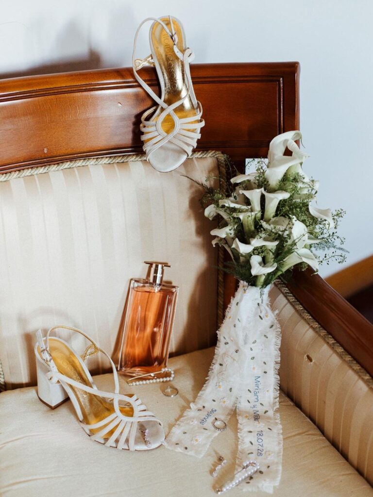 Ramo de calas con cinta bordada personalizada junto a los zapatos y el perfume de la novia