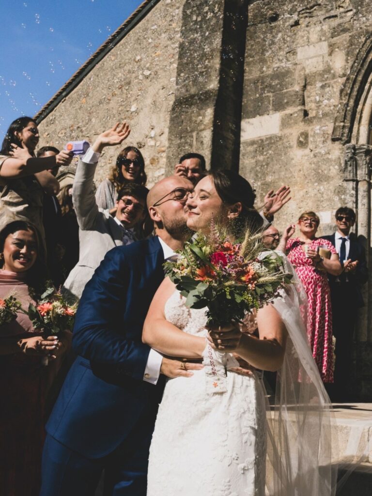 Novios besándose a la salida de la ceremonia, la novia con el ramo y la cinta bordada