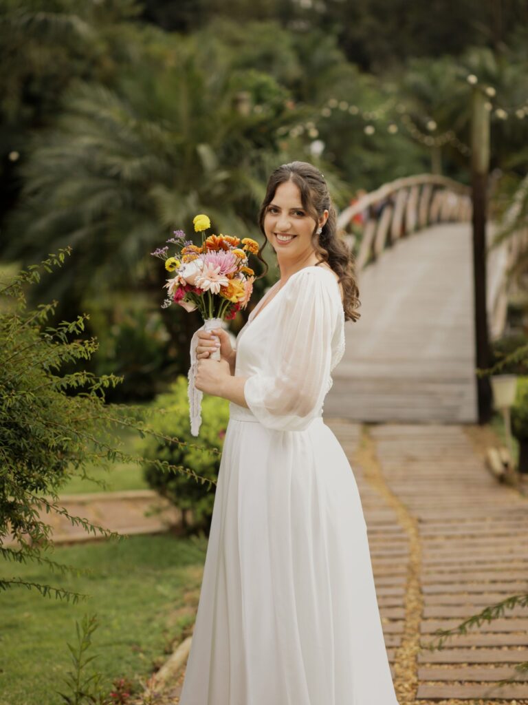 Novia sonriendo con su ramo de flores y la cinta bordada personalizada en un jardín de Brasil
