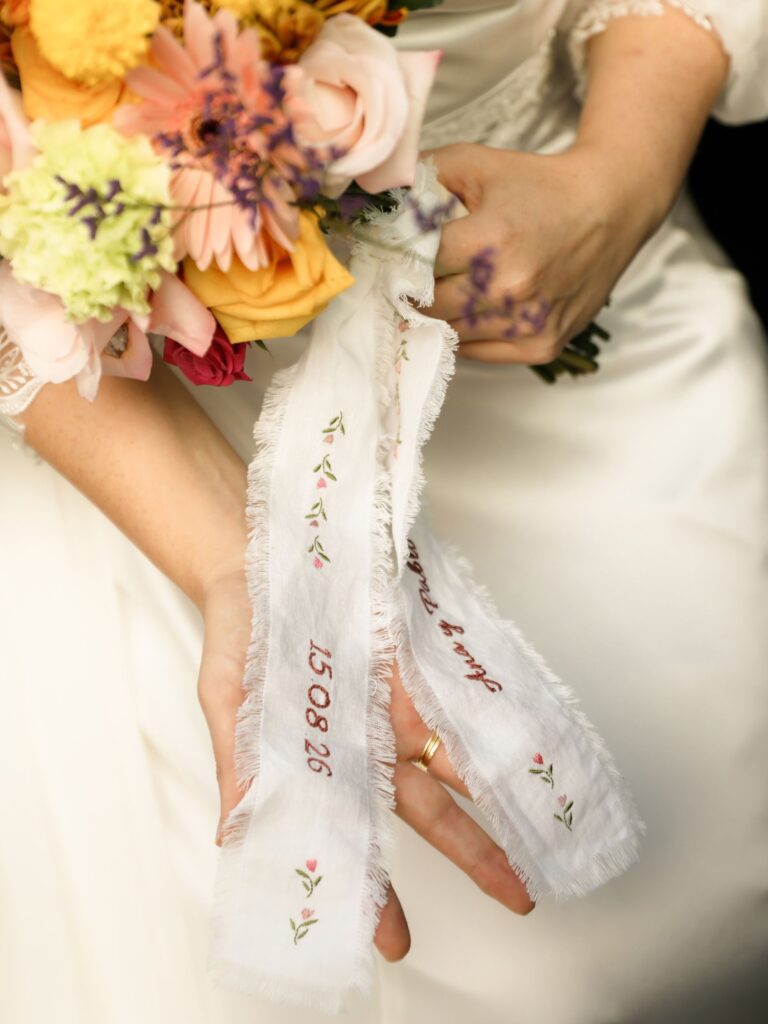 Cinta bordada para ramo de novia con los nombres Ana y Pablo, la fecha 15.08.26 y una cenefa de flores