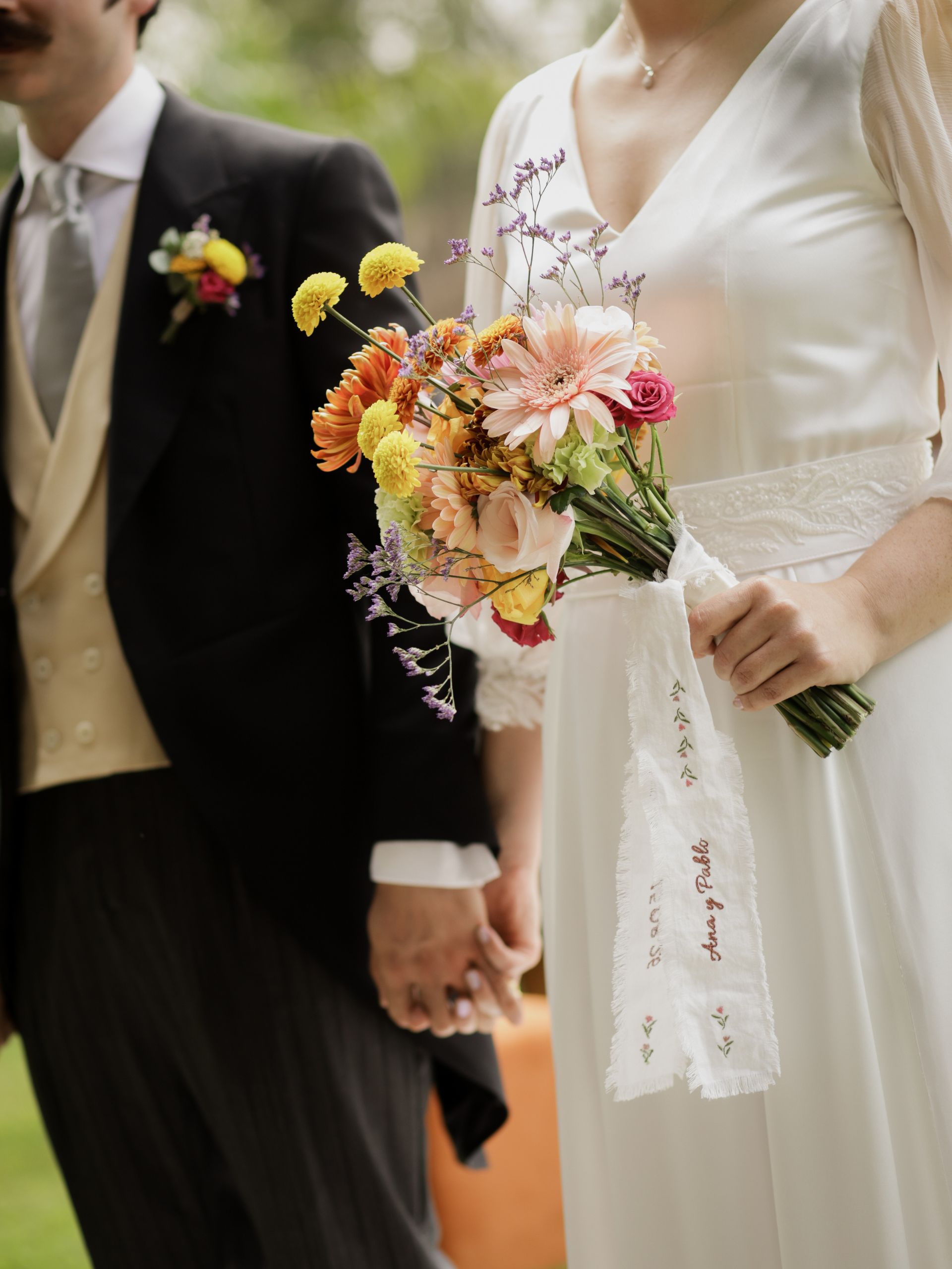 Novios cogidos de la mano, la novia con el ramo y la cinta bordada con sus nombres